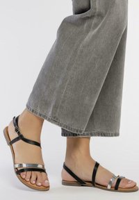 Sandales en cuir noir avec des accents métalliques, semelle plate, bride de cheville réglable et design ouvert, associées à un pantalon ample gris.