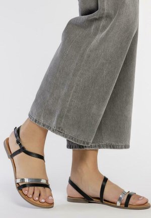 Sandalias de cuero negro con acentos metálicos, suela plana, tira de tobillo ajustable y diseño abierto en la punta, combinadas con pantalones de pierna ancha grises.