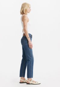 Jeans de mezclilla azul con corte recto y cinturilla de altura media, llevados con una camiseta sin mangas blanca y sandalias blancas con detalles plateados.