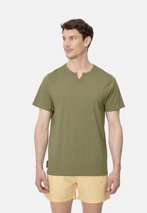 Hombre con camisa henley de manga corta de color verde oliva y pantalones cortos amarillo claro, de pie y mirando hacia un lado sobre un fondo liso.