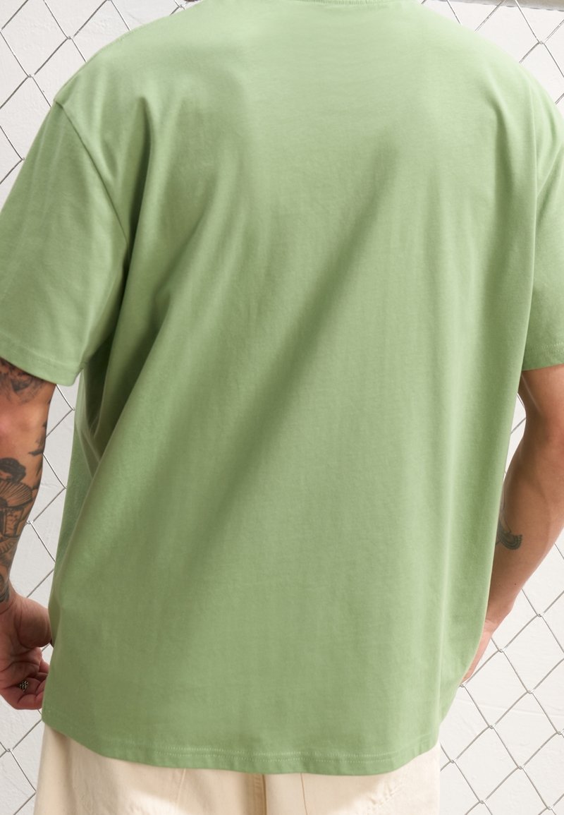 Homme portant une chemise à manches courtes vert clair et un pantalon beige, debout dos à la caméra, montrant des tatouages sur les bras et un fond de clôture en chaîne.