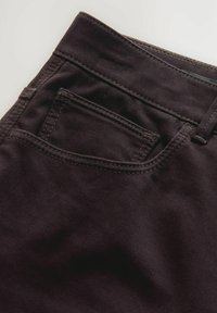 Suur lähikuva mustadest denim-est püksi taskust, kus on nähtav õmblus ja vöökoht heledal taustal.