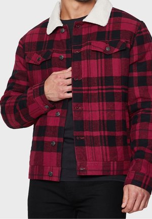 Homme portant une veste à carreaux rouge et noire avec un col en polaire blanche, chemise noire en dessous, ajustant le devant près de la zone de la poitrine.