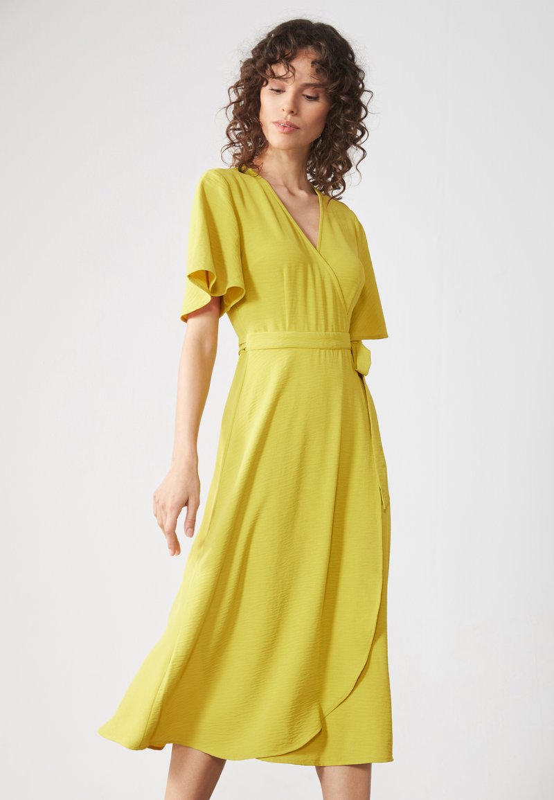 Gelbes Wickelkleid mit kurzen, fließenden Ärmeln, tailliertem Bund und einem schwingenden Saum. Hergestellt aus leichtem Stoff mit strukturierter Oberfläche.