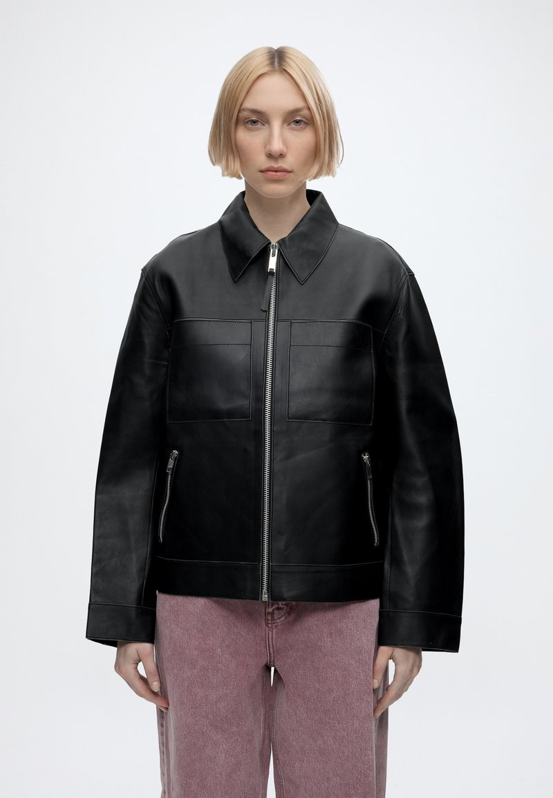 Personne aux cheveux courts et blonds portant une veste en cuir noire zippée avec de grandes poches et un pantalon mauve sur un fond uni.