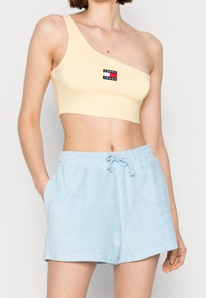 Vrouwelijke torso met een lichtgeel een-shoulder crop top met Tommy Jeans-logo en lichtblauwe badstof shorts met trekkoord.