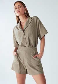 Mujer vistiendo un mono beige de manga corta con botones, grandes bolsillos frontales y pendientes de aro dorados, con las manos en los bolsillos contra un fondo blanco.