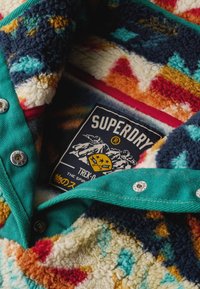 Colorful fleece jacket featuring a multicolored, patterned interior with turquoise accents and silver snap buttons. Includes a branded label.
