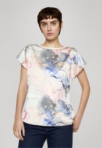 Mujer con cabello corto y oscuro, vistiendo una blusa con patrón de acuarela en tonos pastel y unos vaqueros oscuros, de pie frente a un fondo gris liso.