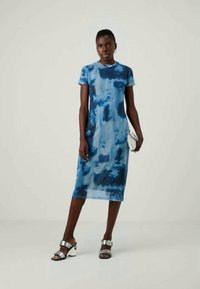 Niebieska sukienka midi w tie-dye z krótkimi rękawami i okrągłym dekoltem, w zestawieniu z srebrnymi sandałami na wysokim obcasie oraz małą białą kopertówką.