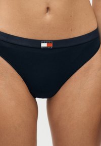 Γυναίκα που φοράει μαύρο κάτω μέρος μαγιό Tommy Hilfiger με το λογότυπο στο μπροστινό μέρος της μέσης.