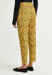 Pantaloni gialli con motivo geometrico che presenta forme marroni e crema alternate, abbinati a scarpe basse nere.