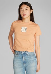 Camiseta de manga corta color durazno hecha de algodón, con un logo cuadrado en negro y crema en el centro. Diseño casual y de corte recto.