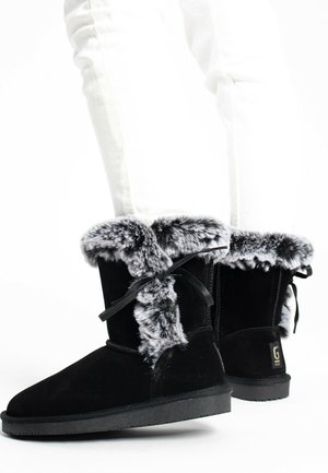 Schwarze Wildlederstiefel mit einem kuscheligen grauen Fellbesatz und Schnürdetail, flacher Gummisohle und einem Logo an der Ferse.
