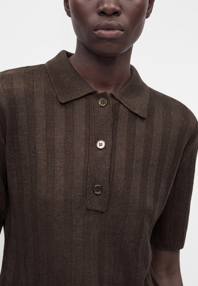 Gros plan d’une personne portant un polo en maille côtelée marron avec un col et trois boutons sur le devant.