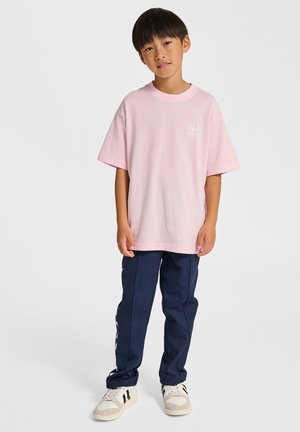 Ljusrosa kortärmad t-shirt med en liten vit logotyp på bröstet, tillsammans med marinblå byxor och beige sneakers.