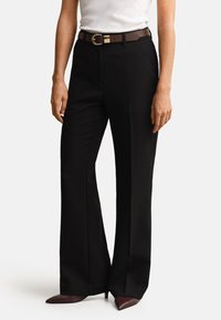 Femeie purtând pantaloni negri drepti cu o curea maro, bluză albă fără mâneci și pantofi cu toc maro și vârf ascuțit pe un fundal simplu.