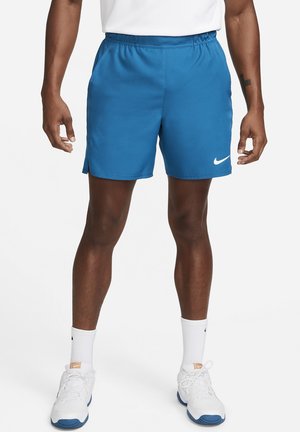 Mann trägt blaue Sportshorts, weiße Socken und weiße Turnschuhe und steht vor einem einfarbigen Hintergrund.