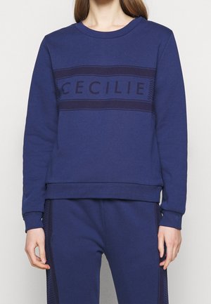 Kvinde iført en mørkeblå sweatshirt med teksten "CECILIE" på brystet og matchende mørkeblå bukser med sidelommer.