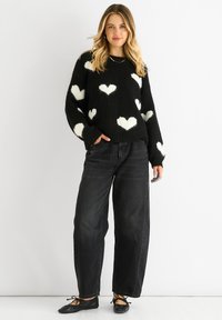 Black knit sweater with white heart patterns, oversized fit, ribbed cuffs, paired with high-waisted black wide-leg jeans and black flats.