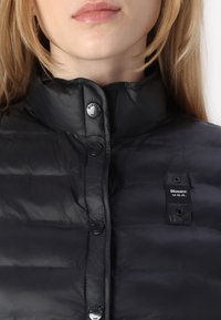 Sort puffer-jakke med høj krave, med trykknapper og et logo-label på venstre bryst. Stoffet har en glat, blank overflade.