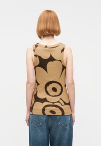  Άτομο με κοντά κόκκινα μαλλιά που φοράει μπεζ αμάνικο τοπ με μεγάλα σκουρόχρωμα καφέ λουλουδένια σχέδια και φαρδιά μπλε τζιν, εμφανιζόμενο από πίσω.