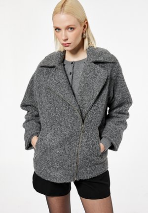 Jonge vrouw draagt een gestructureerd grijs jack met ritssluiting over een grijze top met knopen, zwarte korte broek en creolen, met handen in de jaszakken.