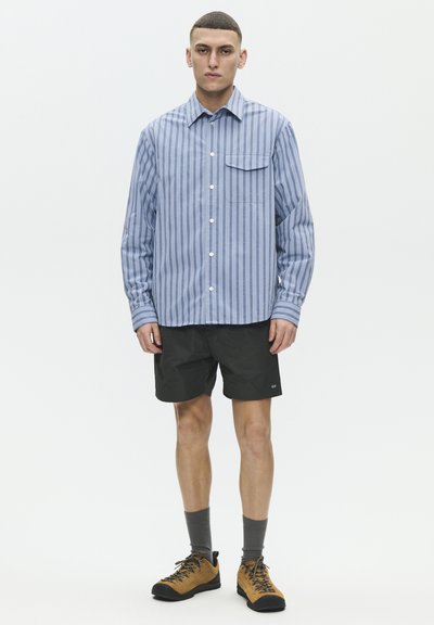 Joven de pie con una camisa de manga larga de rayas azules abotonada, pantalones cortos negros, calcetines grises y zapatos de senderismo marrones sobre un fondo blanco.