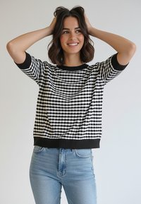 Czarno-biały sweter w kratę z krótkimi rękawami i czarnym prążkowanym wykończeniem. Połączony z jasnoniebieskimi dżinsami.