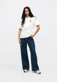 Biela bavlnená tričko s okrúhlym výstrihom a prednou grafikou, skombinované s širokými modrými džínsami. Model nosí biele športové tenisky.