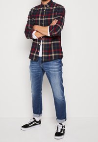 Koszula z flaneli w kratę w kolorach granatowym, czerwonym i beżowym, zestawiona z jasnoniebieskimi podwiniętymi dżinsami oraz czarnymi trampkami z białymi akcentami.