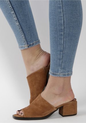 Pieds portant un jean skinny bleu clair et des mules ouvertes en daim marron à talons blocs sur un fond uni.