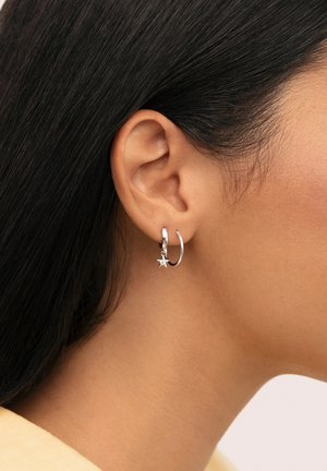 Pendiente de aro de plata con un pequeño charm en forma de estrella colgante. La vista en primer plano muestra un acabado pulido y un diseño sencillo, que realza la oreja.
