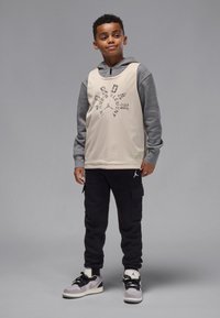 Echipament sportiv pentru băieți, format dintr-un top bej fără mâneci cu glugă din plasă, mâneci gri și pantaloni negri cargo, completat de adidași gri și negri.