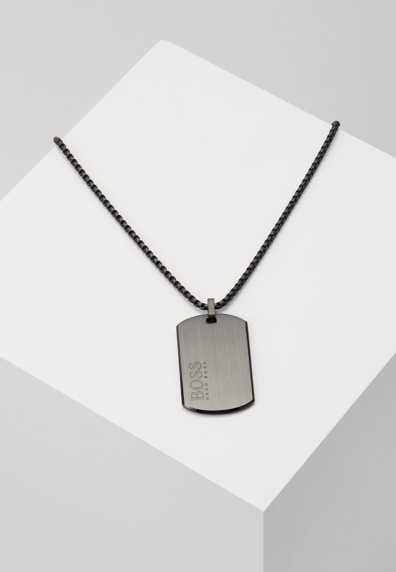 Pendentif plaque militaire en métal noir gravé du logo BOSS Hugo Boss sur une chaîne noire, exposé sur un socle blanc.