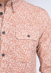 Camisa de manga corta, abotonada, con un estampado floral en naranja y blanco. Presenta dos bolsillos en el pecho y botones negros. Tejido de algodón.