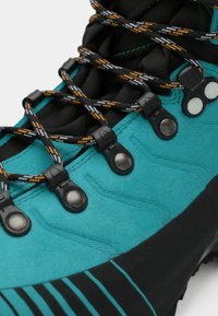 Primeiro plano de uma bota de hiking em verde-azulado com atacadores pretos e laranjas em padrão, passados por ilhós de metal pretos sobre fundo branco.