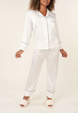 Ensemble de pyjama en satin blanc avec des bordures noires, comprenant un haut à manches longues à boutons et un pantalon droit, porté avec des chaussons blancs duveteux.