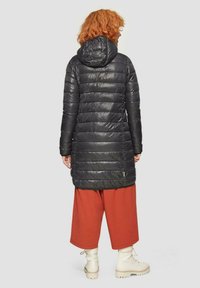 Schwarzer, gepolsterter, knielanger Mantel mit Kapuze. Mit horizontaler Steppung und einem kontrastierenden Akzent. Kombiniert mit orangefarbenen, weit geschnittenen Hosen und cremefarbenen Stiefeln.