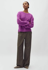 Frau steht mit verschränkten Armen, trägt einen lila gerippten Pullover, weite braune Hosen und spitze braune Schuhe vor einem einfarbigen Hintergrund.