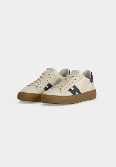 Ζευγάρι μπεζ χαμηλά sneakers με καφέ σόλες, ναυτικά μπλε ακροδάχτυλα και λεπτομέρειες σε ναυτικό μπλε και άσπρο στο πλάι, παρουσιάζονται σε λευκό φόντο.