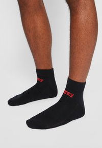 Schwarze Knöchelsocken aus weichem Stoff. Rotes Logo auf der Außenseite aufgedruckt. Verfügt über einen gerippten Bund für einen sicheren Sitz.