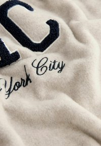 Cremefarvet stof med et tekstureret blåt broderet "C" og "York City" i kursiv. Blød og hyggelig tekstur.