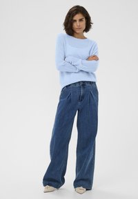 Svetlo modrý pletený sveter s dlhými rukávmi, kombinovaný s voľnými, vysokými tmavomodrými džínsami. Model stojí so založenými rukami.