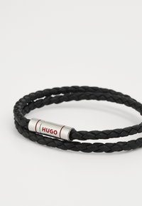 Svart flätad läderarmband med ett silverfärgat lås som har röda detaljer och texten "HUGO" graverad.