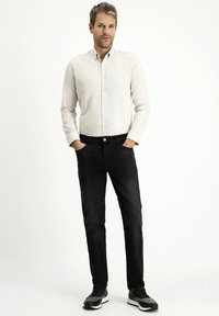Camicia a maniche lunghe di colore chiaro, jeans neri e sneakers nere. La camicia presenta un colletto button-down e un tessuto testurizzato.