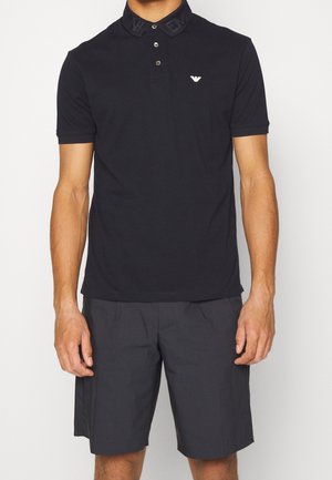 Black polo shirt with a textured collar, button closure, and small logo. Paired with dark gray shorts featuring side pockets.