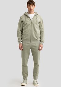 Hellgrüner Kapuzenpullover mit Reißverschluss, Kängurutaschen und Kordelzug. Kombination mit passenden Jogginghosen und Schnürsneakern. Glatte Stofftextur.