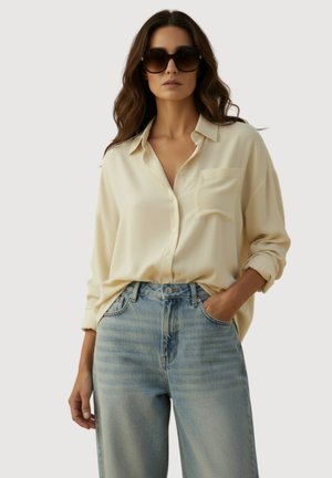 Vrouw draagt een oversized crèmekleurig hemd met knopen, lichtblauwe high-waist jeans en grote donkere zonnebril, poseert met één hand in de zak.