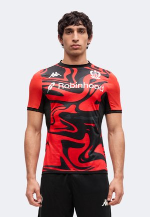 Homme portant un maillot de sport rouge et noir à motif abstrait avec le logo du sponsor Robinhood et un short noir, debout sur un fond uni.